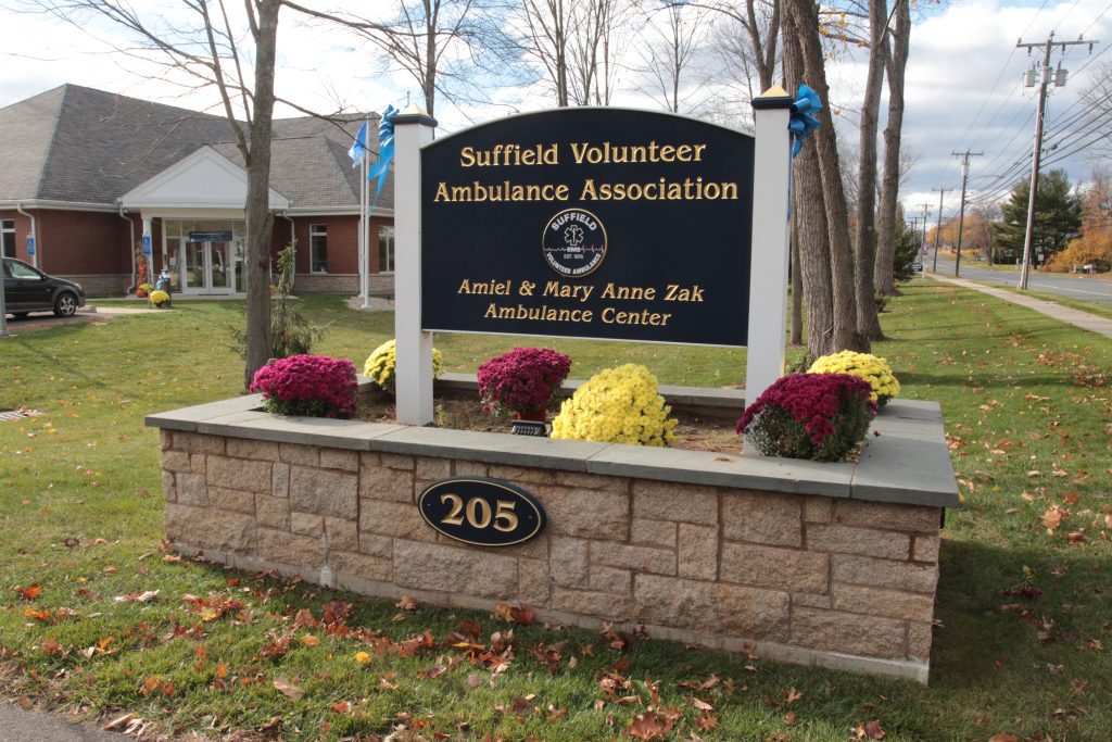 A sign for the Suffield Volunteer Ambulance Association with "Amiel & Mary Anne Zak Ambulance Center" written on it, surrounded by flowers. The sign is located at 205, in front of a brick building and near a road lined with trees.