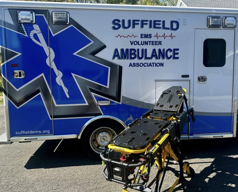 A Suffield EMS Volunteer Ambulance Association vehicle is parked with the back doors closed. A medical stretcher is positioned in front of it on the pavement. The ambulance is primarily white with blue graphics and text.